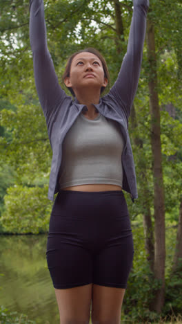 Vertical-Video-Of-Woman-Wearing-Sports-Clothing-Doing-Yoga-Stretches-On-Mat-In-Forest-By-Lake-Or-River-Enjoying-Peace-And-Beauty-Of-Nature-Shot-In-Real-Time-1